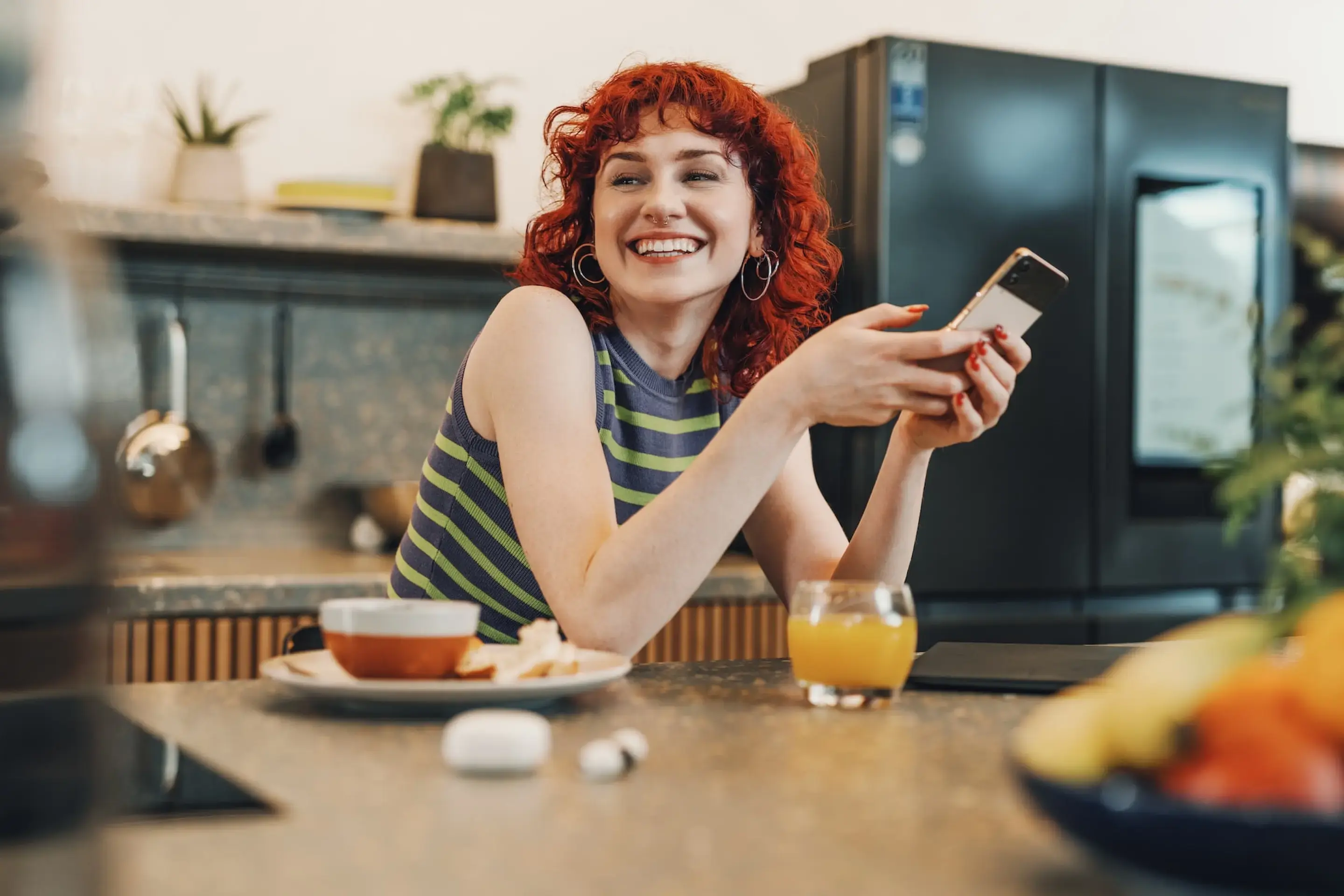 Eine lächelnde Frau sitzt in einer Küche, hält ein Smartphone in der Hand und schaut zur Seite. Vor ihr stehen ein Glas Orangensaft, eine Schale mit Essen und ein Paar kabellose Ohrhörer.