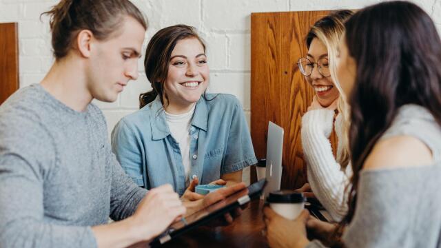 Vier junge Erwachsene sitzen an einem Holztisch in einem gemütlichen Café oder Arbeitsraum. Sie unterhalten sich fröhlich, lachen und arbeiten gemeinsam an einem Tablet und Laptop. Eine entspannte und kreative Atmosphäre ist spürbar.