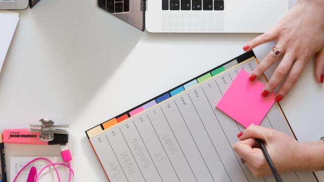 Eine Person sitzt an einem Schreibtisch mit einem Laptop, schreibt auf einen pinkfarbenen Notizzettel und hat eine Planungsmappe mit verschiedenen farbigen Registerkarten vor sich. Auf dem Tisch liegen außerdem Büroartikel wie ein Textmarker, Kopfhörer und ein Stift.
