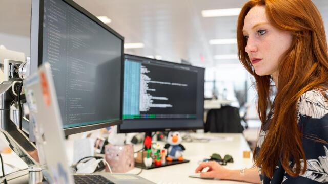 Eine Frau arbeitet an einem Schreibtisch mit mehreren Monitoren, auf denen Programmiercode angezeigt wird. Sie konzentriert sich auf ihre Arbeit in einem modernen Büro.