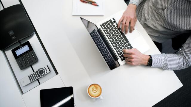 Ein Mann arbeitet an einem Laptop an einem modernen Schreibtisch. Neben ihm stehen eine Tasse Kaffee, ein Tablet, ein Telefon und einige Stifte auf einem Notizblock.